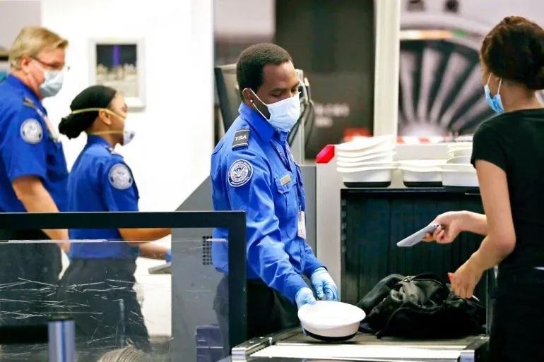 موظفان من إدارة أمن النقل (TSA) يرتديان زيًا رسميًا ويقومون بفحص الأمتعة في مطار، مع وجود مسافرة تتفاعل معهم.