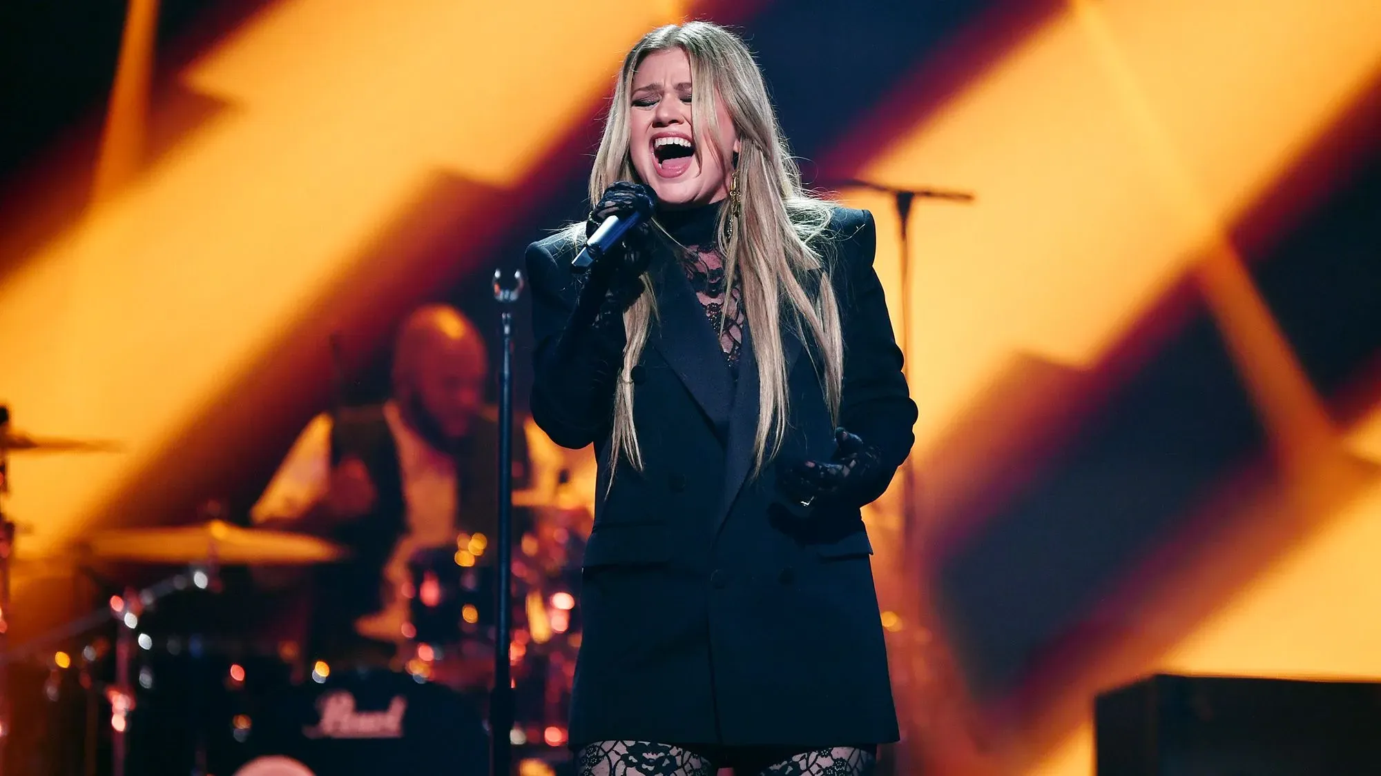 كيلي كلاركسون تؤدي على المسرح، مرتدية ملابس سوداء، مع تعبيرات وجه تعكس مشاعرها بعد تأجيل عرضها في لاس فيغاس.