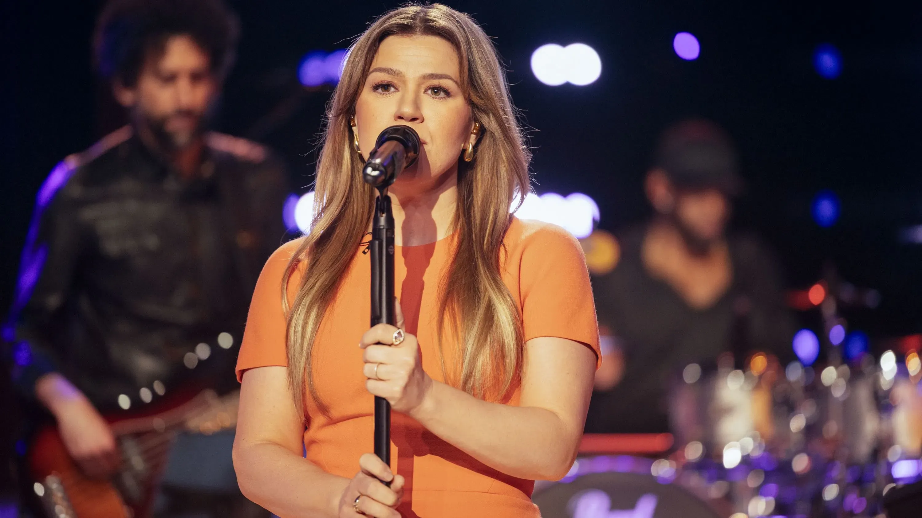 كيلي كلاركسون تغني في الحلقة رقم 1000 من برنامجها الحواري، مع فرقة موسيقية خلفها، تحت أضواء ملونة تعكس أجواء الاحتفال.