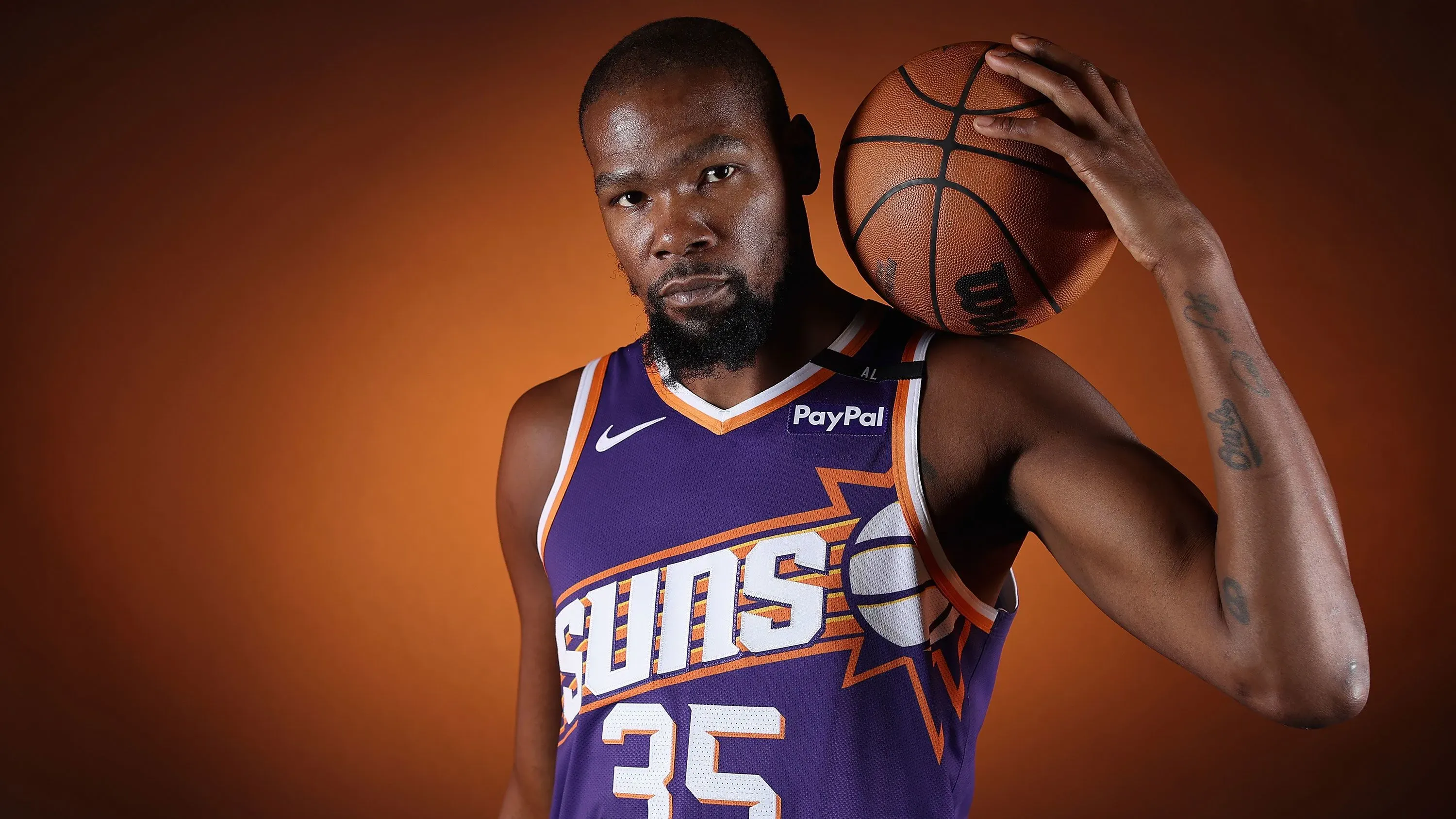 كيفن دورانت، لاعب كرة السلة الشهير، يرتدي زي فريق فينيكس صنز، ممسكًا بكرة سلة، مع خلفية برتقالية تعكس طاقة الدوري الأمريكي للمحترفين.
