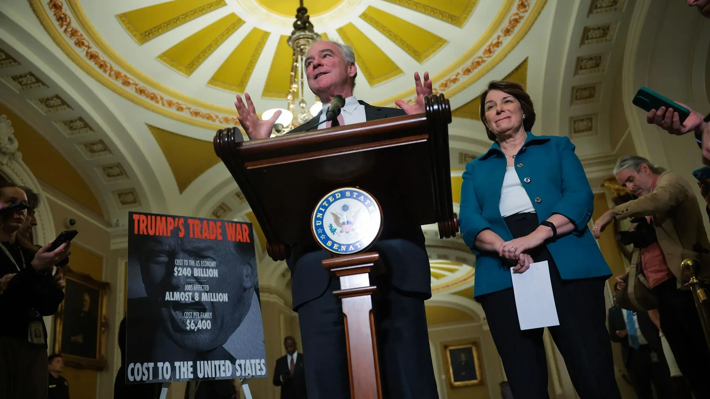 السيناتور تيم كين يتحدث في مجلس الشيوخ عن تأثير الحرب التجارية للرئيس ترامب، مع عرض معلومات مالية على لوحة خلفه.