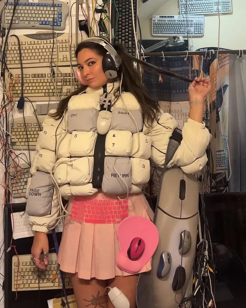 امرأة ترتدي سترة مبطنة مزينة بأزرار لوحة مفاتيح، مع تنورة قصيرة، محاطة بأسلاك وأجهزة كمبيوتر، تعكس أسلوب الموضة المتطرفة.