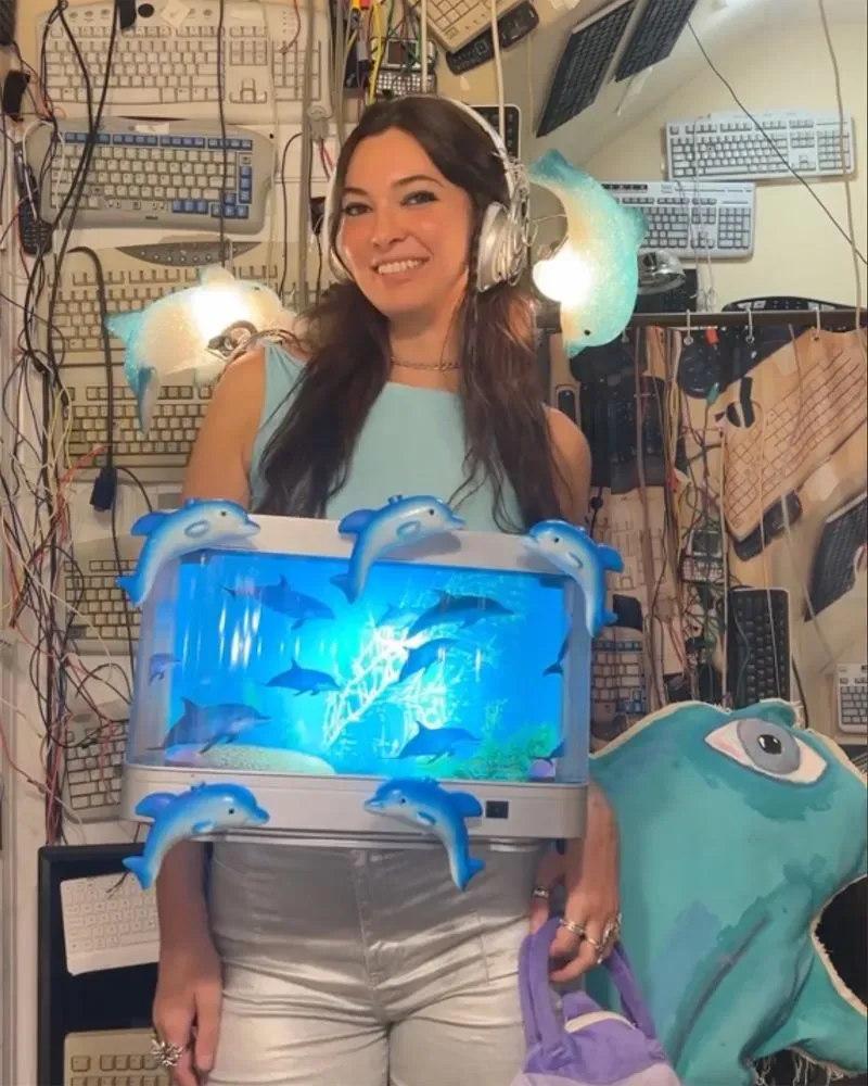 امرأة ترتدي ملابس عصرية، تحمل حوض سمك مزيف مع أسماك ودلافين، محاطة بخلفية من لوحات مفاتيح وأضواء، تعكس أسلوب الموضة المتطرفة.