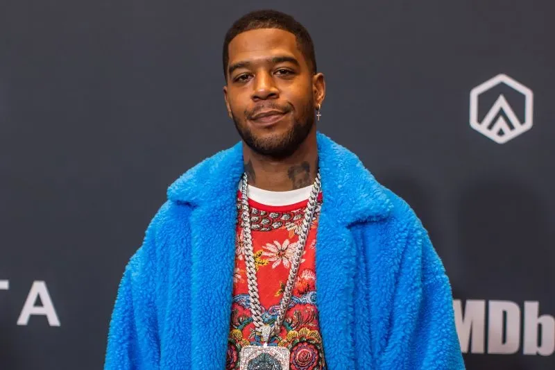 كيد كودي، مغني الراب المعروف، يرتدي سترة زرقاء زاهية ويبتسم أمام خلفية داكنة، مما يعكس شخصيته الفنية الفريدة.