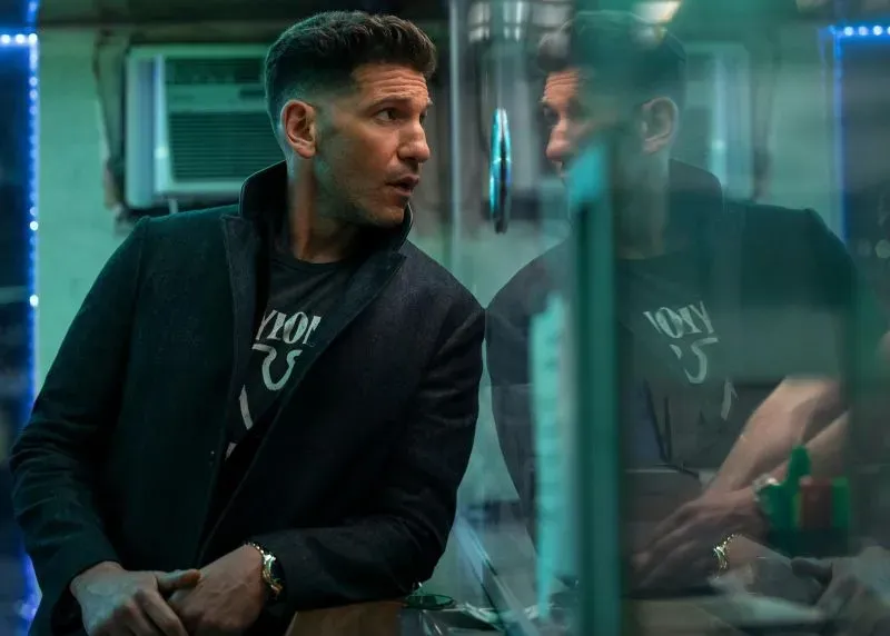 لقطة لرجل يرتدي سترة داكنة، يجلس في مكان داخلي مضاء بأضواء زرقاء، يبدو عليه التفكير العميق، مما يعكس أجواء الإثارة والغموض في الأفلام المرتقبة.