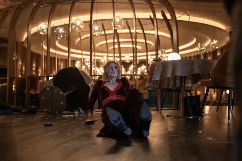 امرأة ترتدي فستانًا أحمر تجلس على الأرض في مطعم فاخر، محاطة بالفوضى والزجاج المكسور، مما يبرز أجواء التوتر في الفيلم.