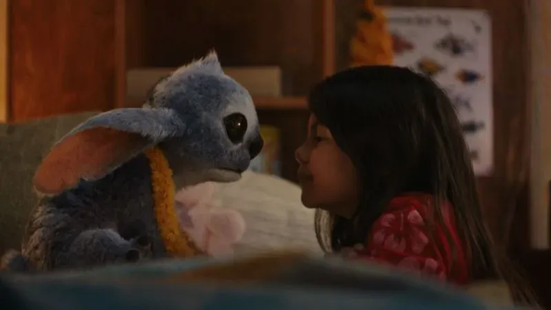 فتاة صغيرة تقترب من دمية زرقاء تشبه كائن فضائي، تعبيرات وجههما تعكس لحظة حميمية ومؤثرة.