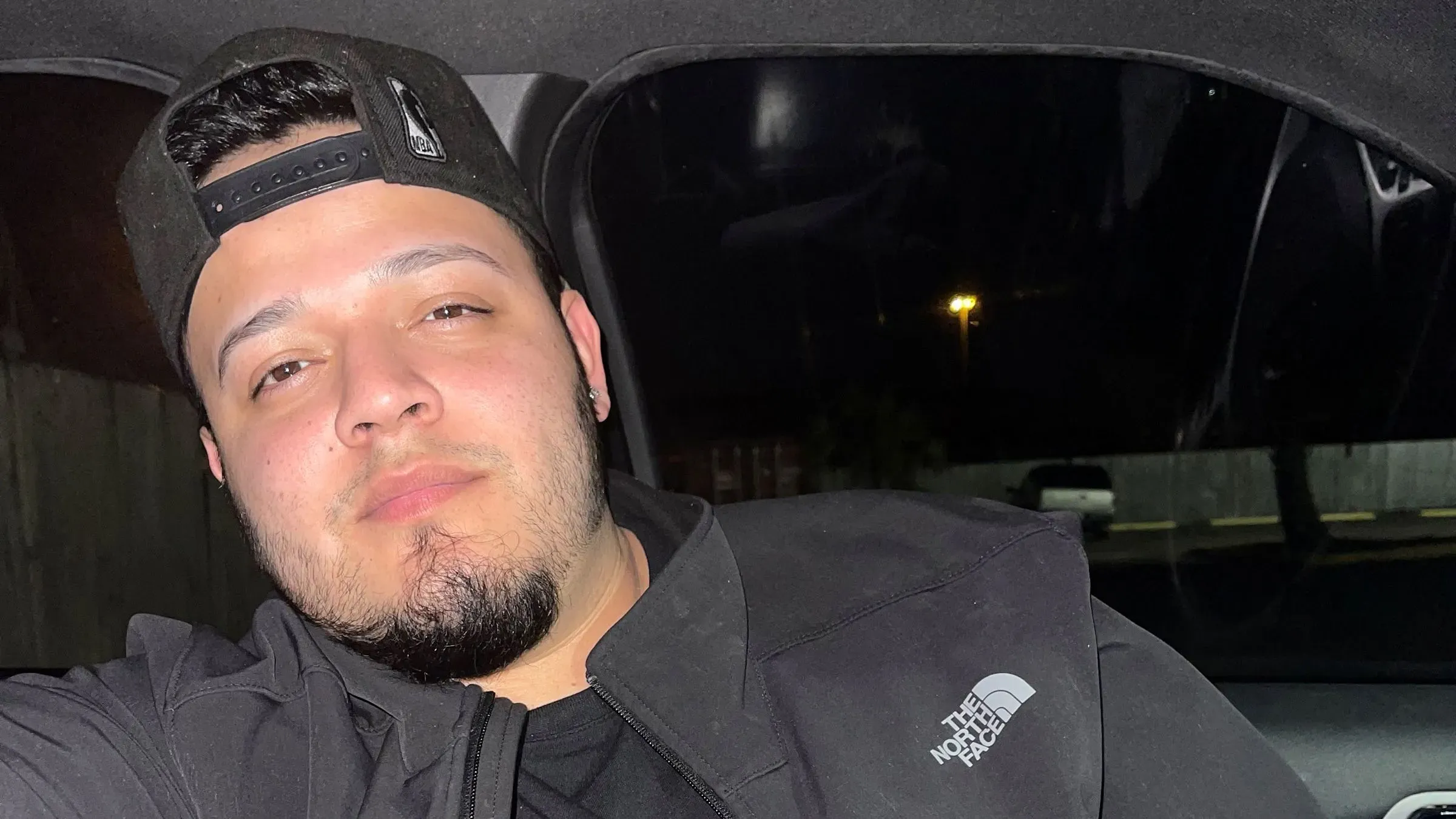 صورة لشاب يجلس في سيارة، يرتدي قبعة سوداء وقميصًا أسود، ويبدو عليه الاسترخاء. تعكس الصورة قضيته القانونية المعقدة مع إدارة ترامب.