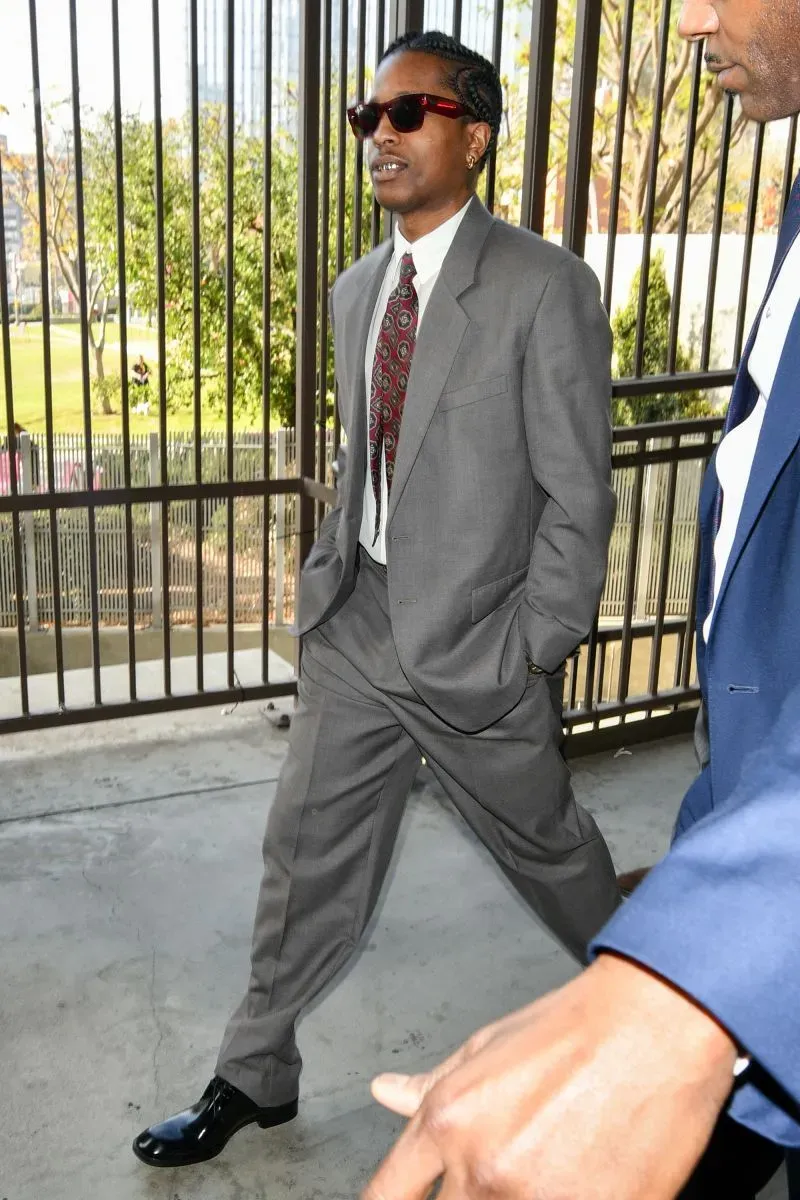 رجل يرتدي بدلة رمادية أنيقة ونظارات شمسية، يسير نحو قاعة المحكمة في باريس، مما يعكس أسلوبه الواثق في سياق محاكمة مشهورة.