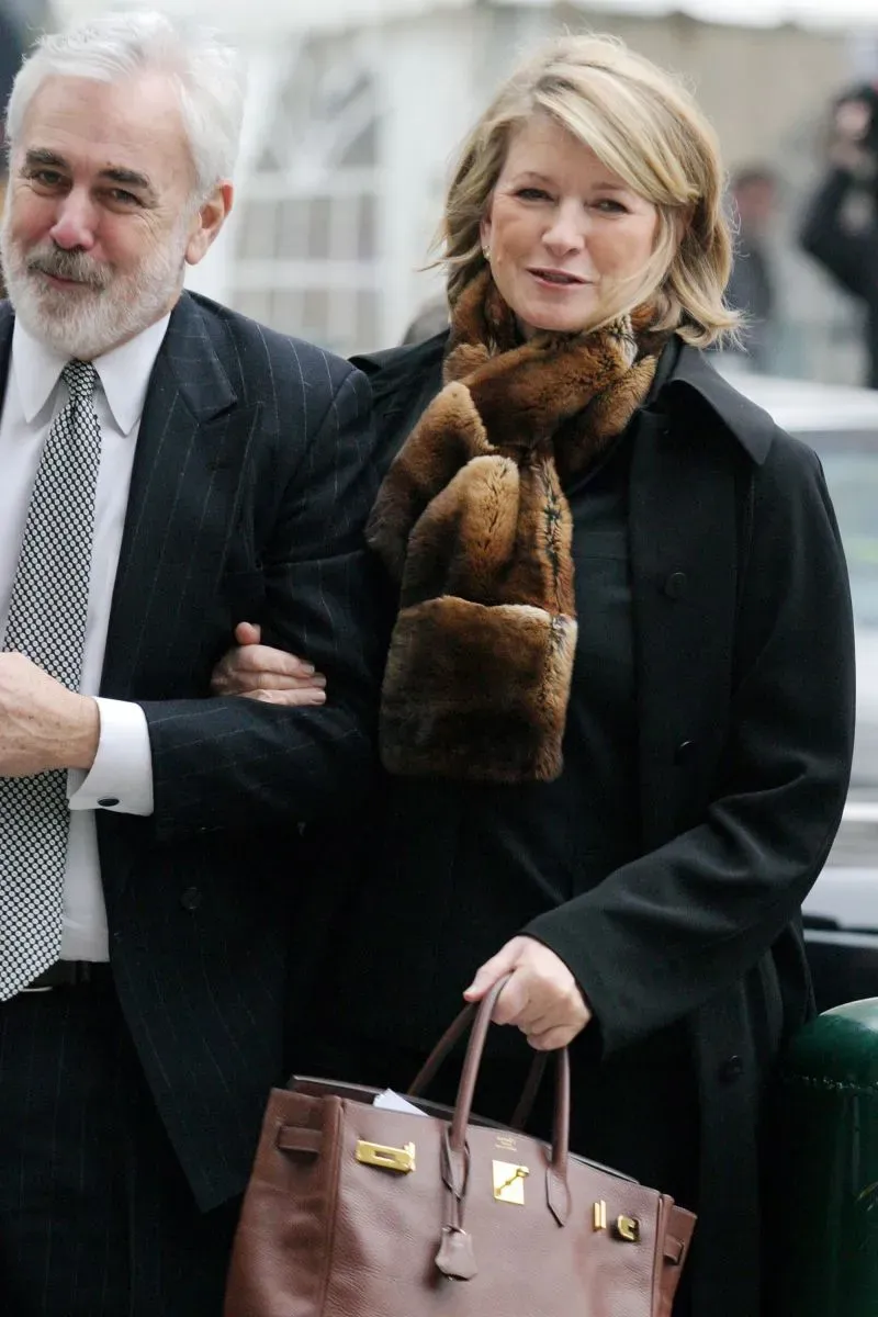 امرأة تحمل حقيبة فاخرة ترتدي سترة سوداء ووشاحًا من الفرو، تسير مع رجل في بدلة، في أجواء محكمة باريسية.