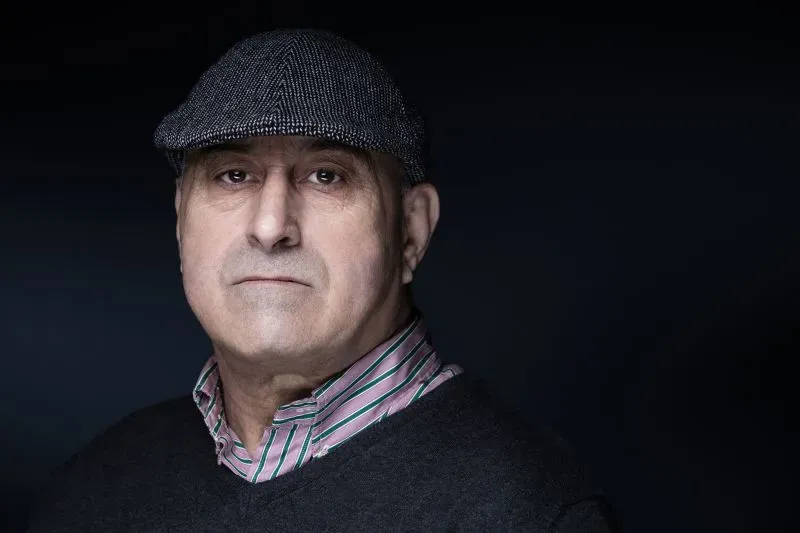 صورة لرجل مسن يرتدي قبعة رمادية وقميص مخطط، يظهر تعبيره الجاد في إضاءة خافتة، مما يعكس أجواء الجريمة في باريس.