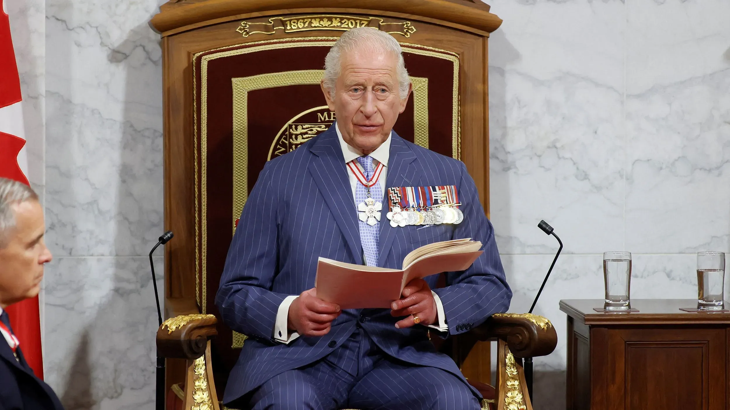 الملك تشارلز الثالث يلقي خطابًا في البرلمان الكندي، مرتديًا بدلة رسمية، ويقرأ من كتاب، مع التركيز على سيادة كندا.