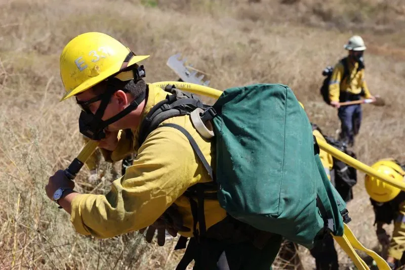 رجال إطفاء يرتدون خوذات وقائية وأقنعة، يحملون خرطوم مياه في منطقة جافة، أثناء مكافحة حرائق الغابات في لوس أنجلوس.