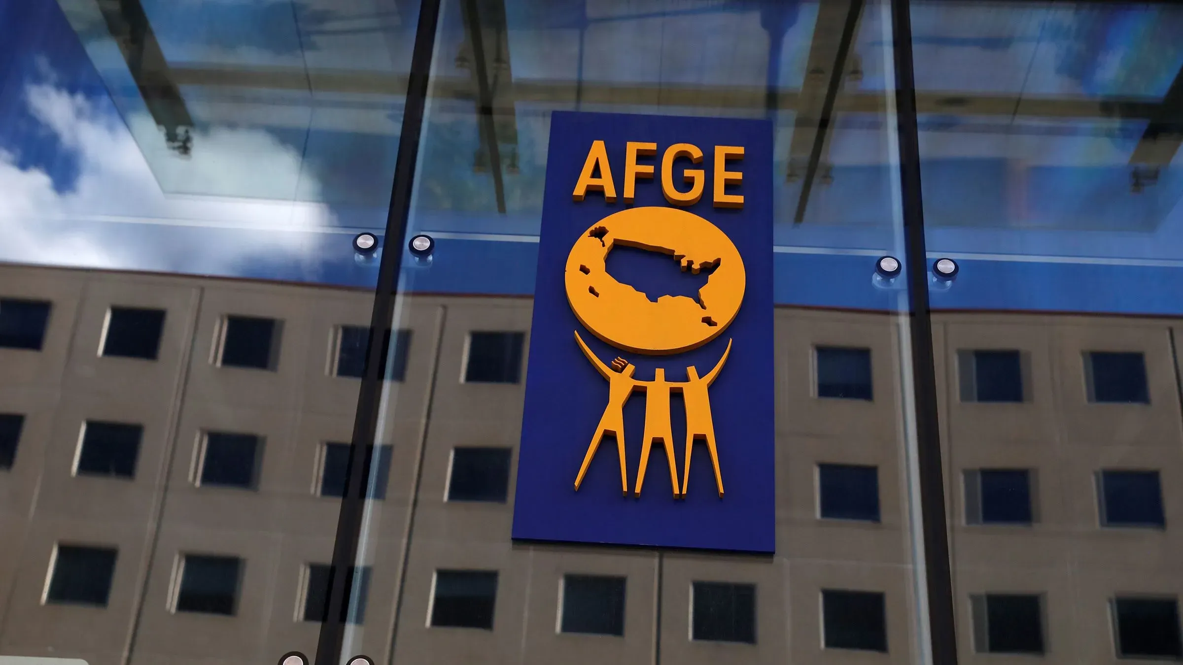 شعار نقابة AFGE يظهر على واجهة زجاجية، مع خلفية لمبنى إداري، يعكس التحديات التي تواجه النقابة في ظل تقليص عدد الموظفين الفيدراليين.