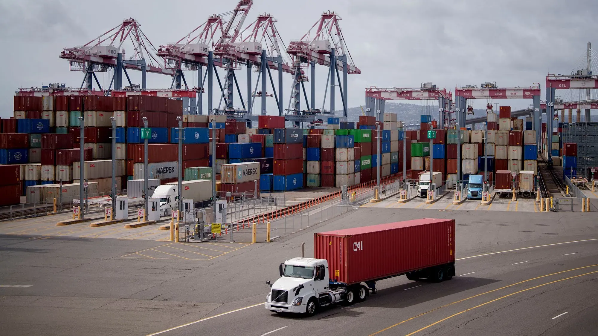 صورة لميناء يحتوي على حاويات شحن ملونة وشاحنة تنقل حاوية، تعكس التوترات التجارية وتأثير سياسات ترامب على الاقتصاد الأمريكي.