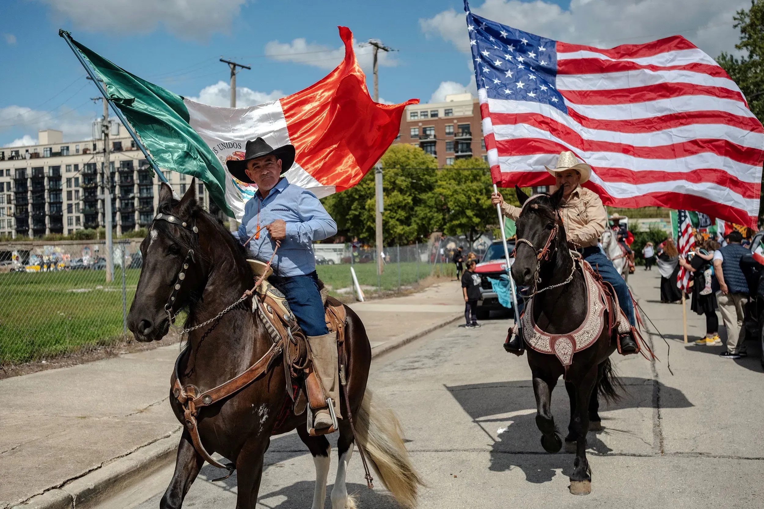 رجال يرتدون ملابس تقليدية يمتطون خيولًا، حاملين علمي المكسيك والولايات المتحدة خلال احتفالات يوم الاستقلال المكسيكي في الشارع.