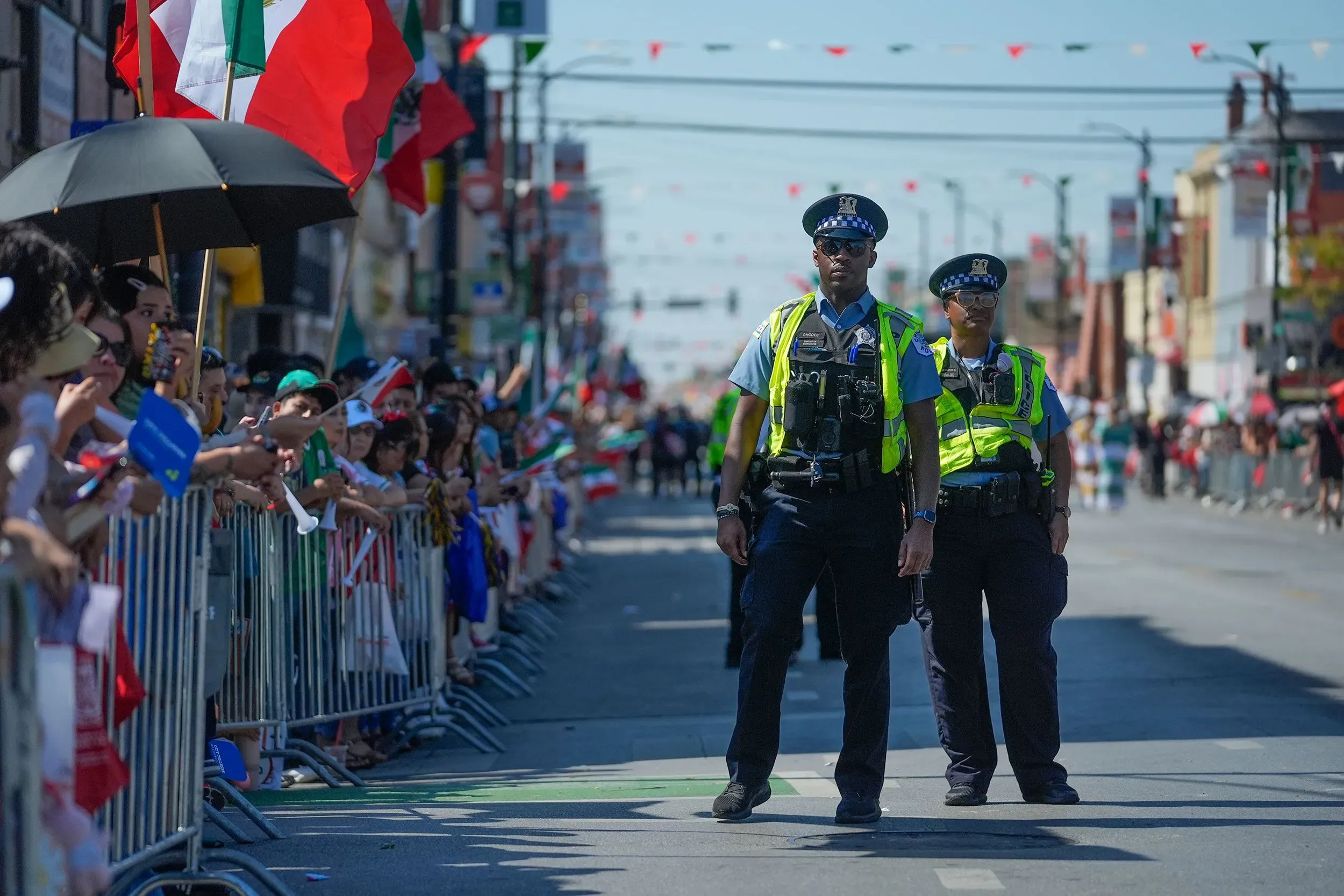 رجال شرطة يرتدون زيهم الرسمي يقفون في مقدمة موكب احتفالي، بينما يحتشد المتفرجون حاملين أعلام المكسيك في الشوارع.