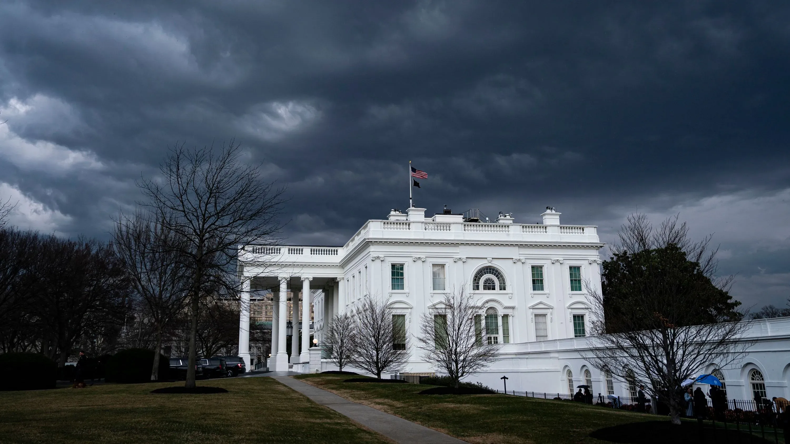 مبنى البيت الأبيض تحت سماء غائمة، مع العلم الأمريكي يرفرف فوقه. الصورة تعكس الأجواء المشحونة حول قضايا الهجرة.
