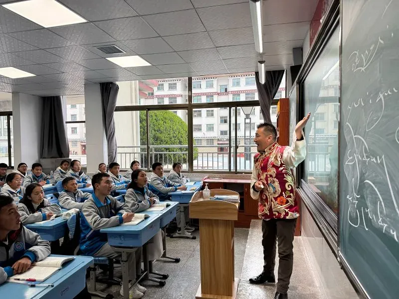 فصل دراسي في التبت يظهر معلمًا يرتدي زيًا تقليديًا يتحدث أمام طلاب يجلسون في صفوف، مع نافذة تطل على مشهد حضري.