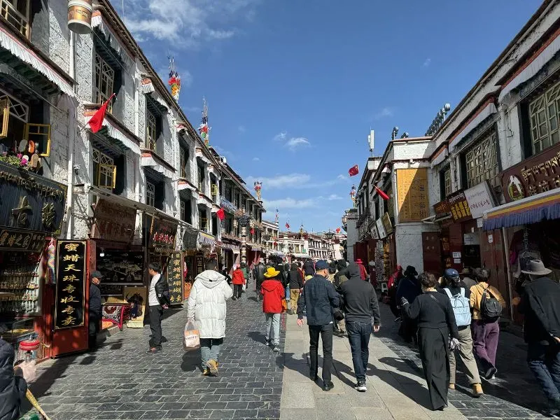 شارع مزدحم في لاسا، التبت، يضم مباني تقليدية مزينة بأعلام وصور، مع مجموعة من الناس يرتدون ملابس متنوعة تحت سماء زرقاء.