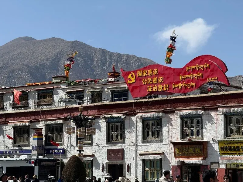 لافتة حمراء تحمل شعارات الحزب الشيوعي الصيني فوق مبنى تقليدي في لاسا، مع جبال خلفية، تعكس التوترات السياسية في التبت.