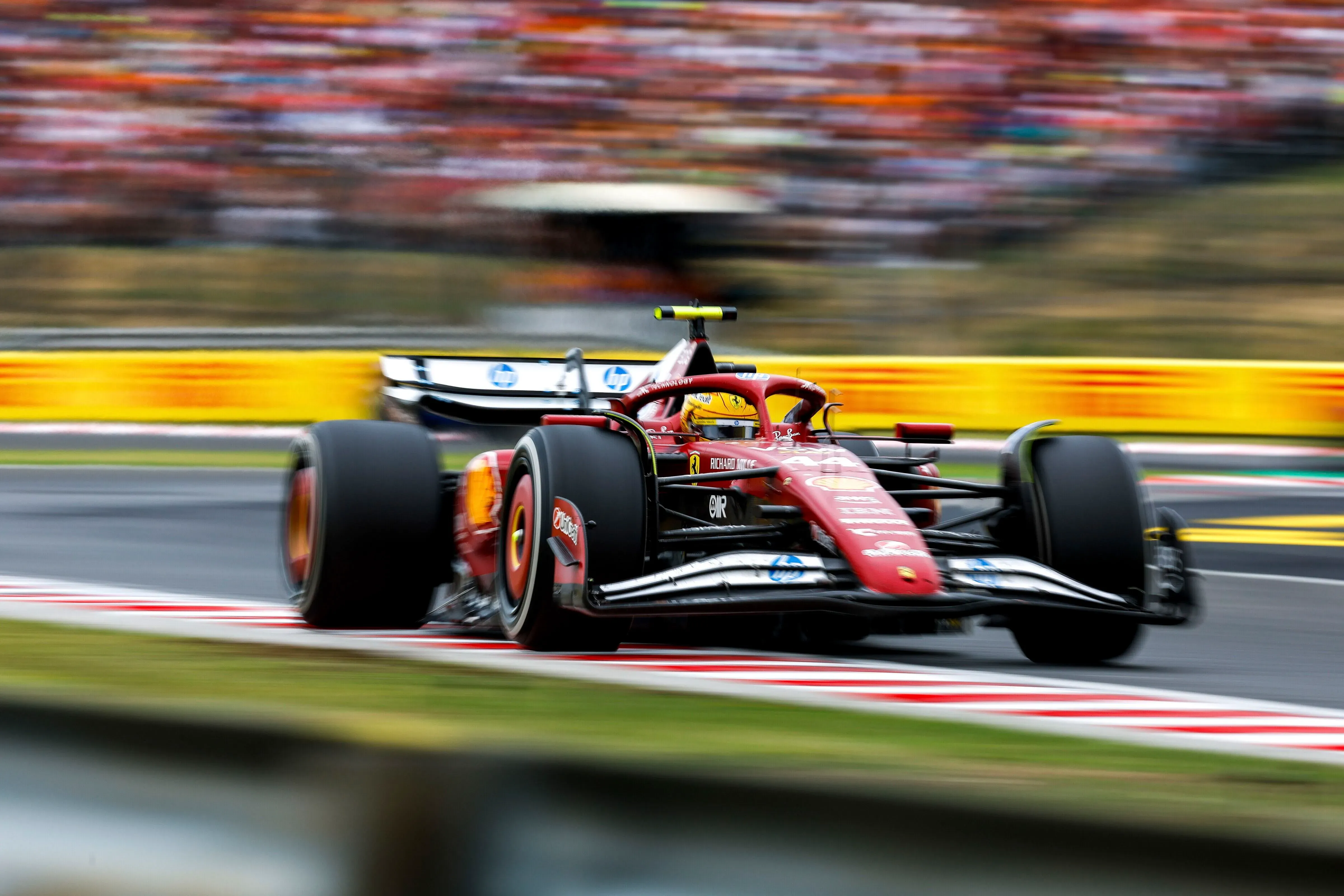 سيارة فيراري تسير بسرعة عالية على حلبة سباق الجائزة الكبرى المجري، مع خلفية جماهيرية ملونة، تعكس تحديات لويس هاميلتون في السباق.