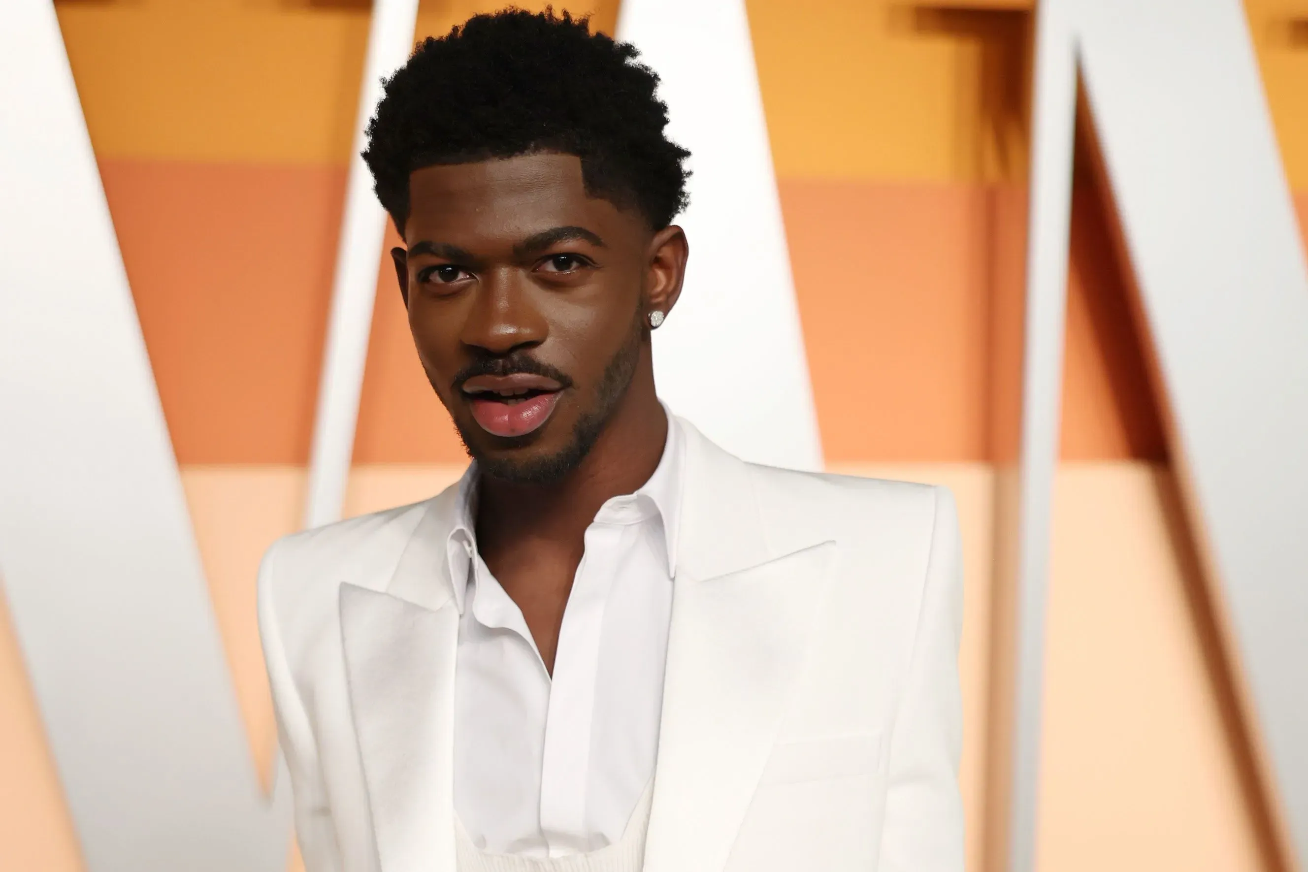 مغني الراب ليل ناس إكس يرتدي بدلة بيضاء ويظهر بابتسامة، في حدث رسمي، مع خلفية ملونة تعكس أسلوبه الفني المبتكر.