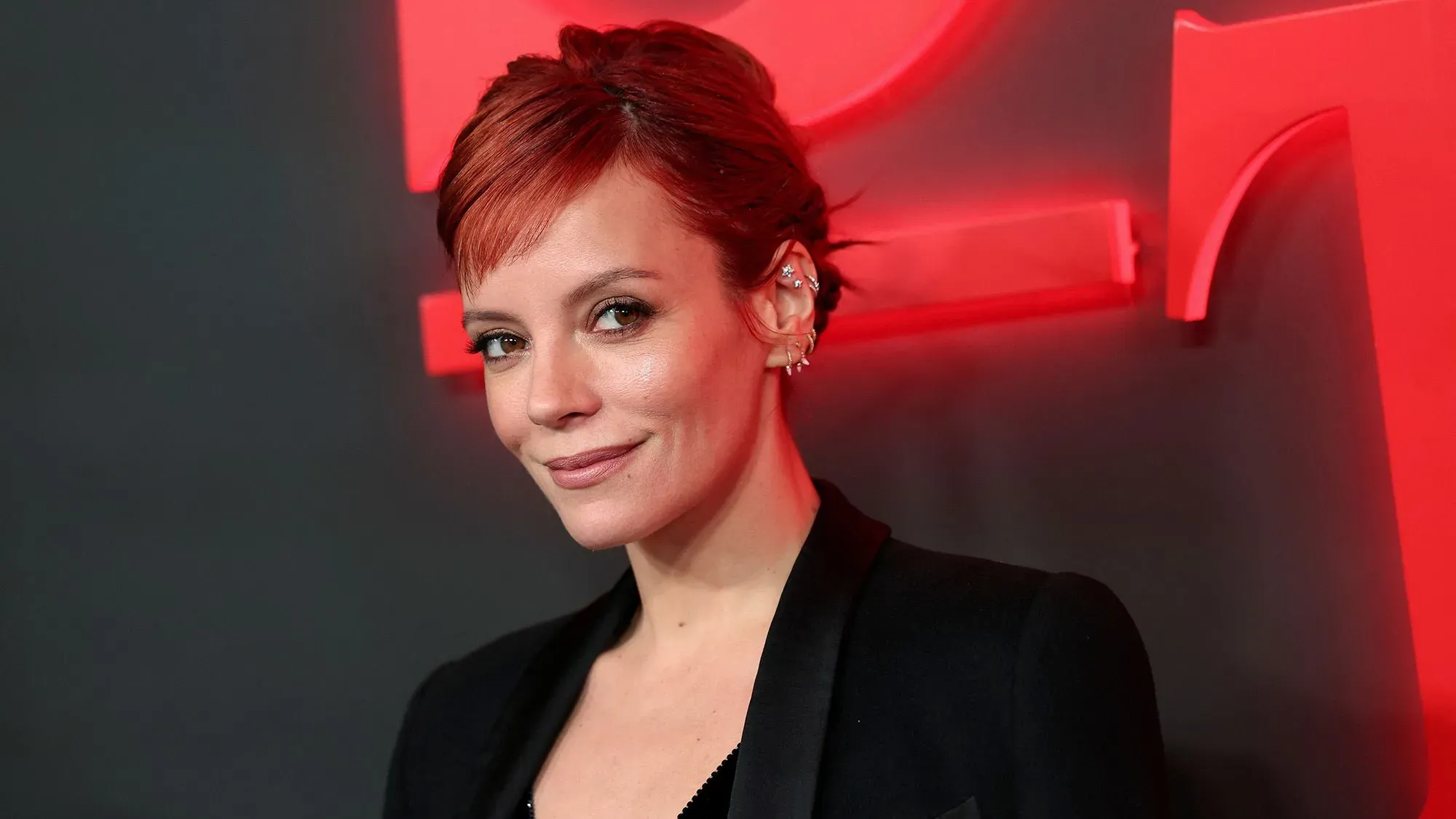 ليلي ألين، نجمة البوب البريطانية، تتحدث في حدث عام، مع خلفية حمراء، تعبيرها يظهر الندم على انتقادها لكاتي بيري.