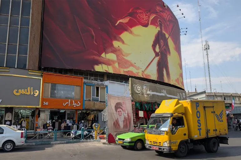 شاحنة بريد إيرانية صفراء تمر أمام مبانٍ تجارية في طهران تحت لوحة جدارية كبيرة تعكس الأوضاع الاقتصادية الصعبة في إيران.