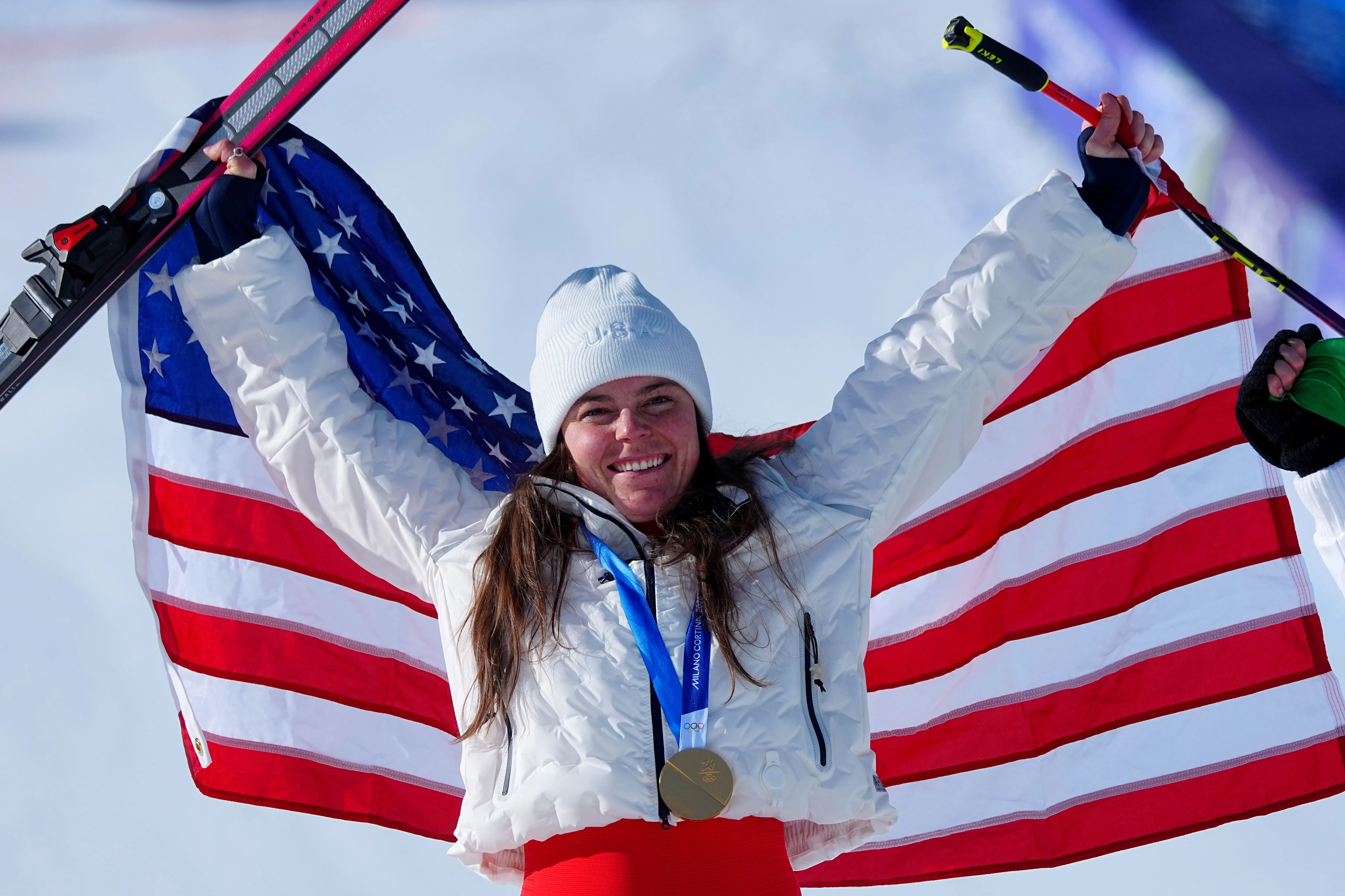 امرأة تحمل علم الولايات المتحدة بفخر، ترتدي ملابس شتوية، وتبتسم بعد تحقيق إنجاز رياضي، مع ميدالية ذهبية حول عنقها.