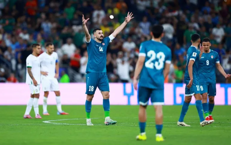أيمن حسين، مهاجم المنتخب العراقي، يحتفل بعد تسجيل هدف في مباراة كأس العالم، محاطًا بزملائه في الفريق، مع جمهور متحمس في الخلفية.