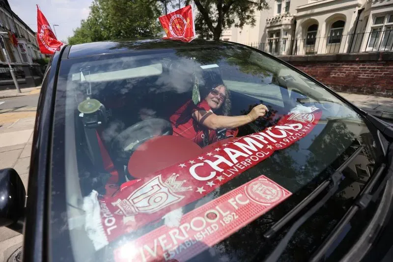 امرأة ترتدي ملابس ليفربول تحتفل بفوز فريقها بلقب الدوري، تجلس في سيارة مزينة بأعلام وشعارات النادي، تعبيراً عن الفخر والفرحة.