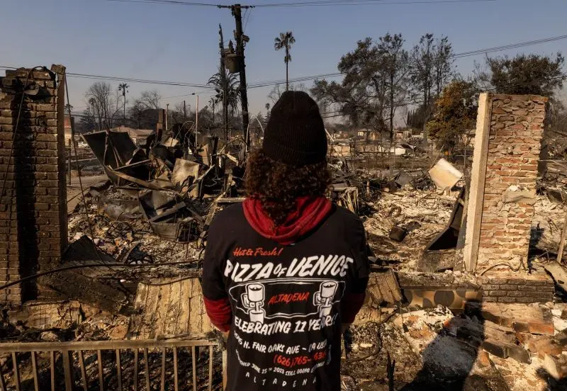 رجل يقف أمام أنقاض مطعم مدمر بعد حريق إيتون، بملابس تحمل شعار "بيتزا فينيس"، حيث خطر العواصف يلقي بظلاله على المنطقة.