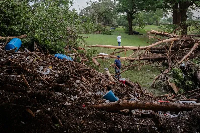تظهر الصورة شخصين يتجولان بين الحطام والأشجار المتساقطة في منطقة تضررت من الفيضانات في وسط تكساس، مما يعكس آثار الكارثة.