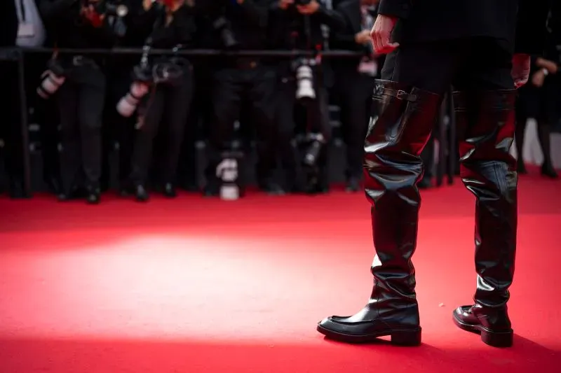 ألكسندر سكارسغارد يرتدي أحذية جلدية طويلة تصل إلى الفخذ في مهرجان كان السينمائي، محاطًا بعدسات الكاميرات، مما يبرز تطور الموضة الجريئة.