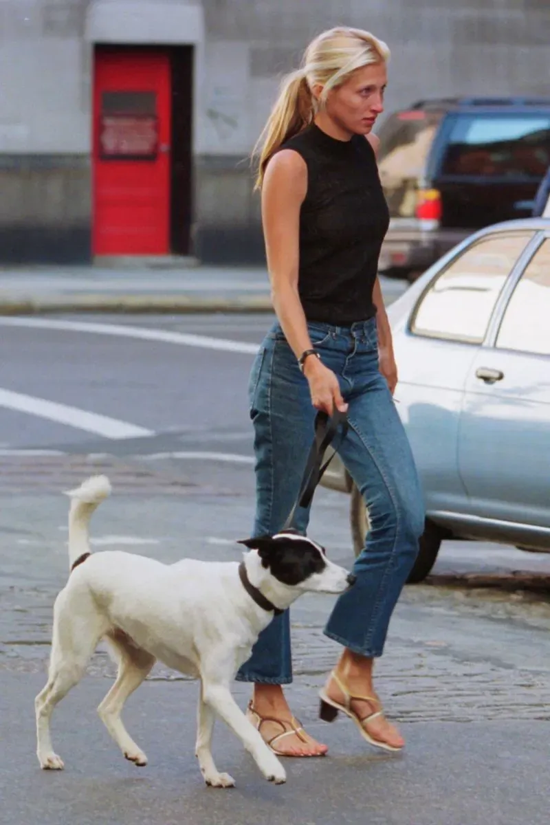 امرأة تسير في الشارع مع كلبها، ترتدي قميصًا أسود وسروال جينز، تعكس أسلوب كارولين بيسيت-كنيدي الأنيق.