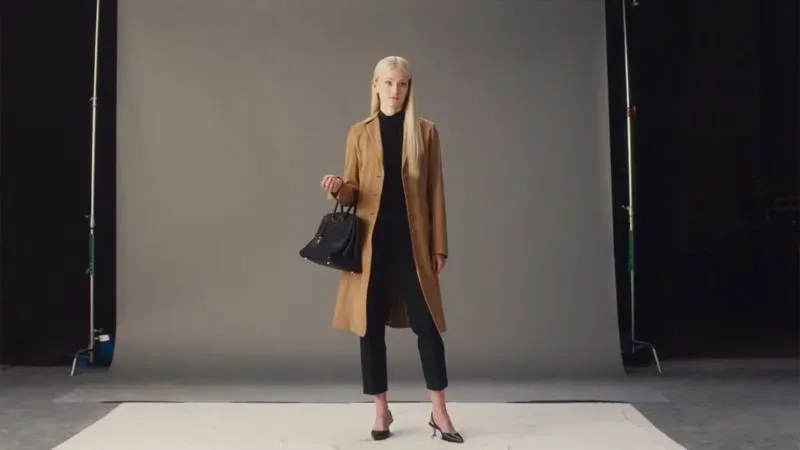 امرأة ترتدي معطفًا جملياً وحقيبة هيرميس بيركين، تقف على خلفية رمادية، تعكس أسلوب كارولين بيسيت-كنيدي.