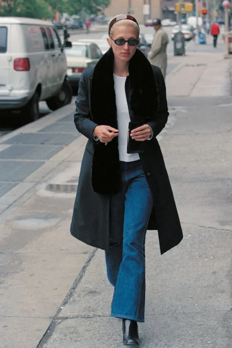 امرأة ترتدي معطفًا أسود طويلًا مع وشاح أسود، وتحتسي مشروبًا أثناء سيرها في الشارع. تعكس إطلالتها أسلوب كارولين بيسيت-كنيدي الأنيق.