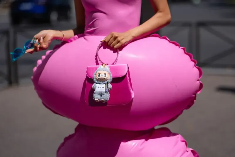 امرأة ترتدي ملابس وردية وتحمل حقيبة صغيرة وردية مزينة بلعبة لابوبو، بينما تقف أمام خلفية حضرية.