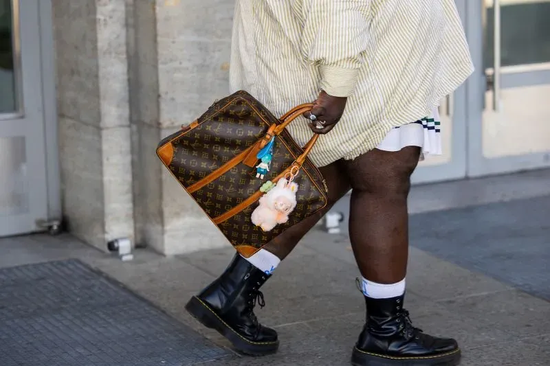 شخص يحمل حقيبة لويس فويتون مزينة بقطع ملونة، يرتدي قميصًا مخططًا وسروالًا قصيرًا، بينما يمشي أمام مكتبة نيويورك العامة.