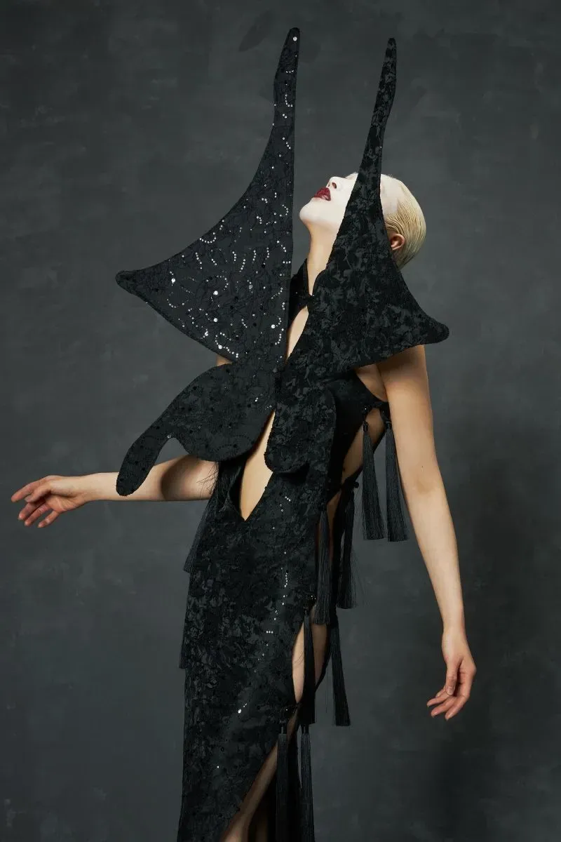 فستان أسود دراماتيكي بتصميم مبتكر من علامة كواي لي، مزين بزخارف فنية، يعكس التأثيرات الثقافية الآسيوية، مع تفاصيل جريئة.