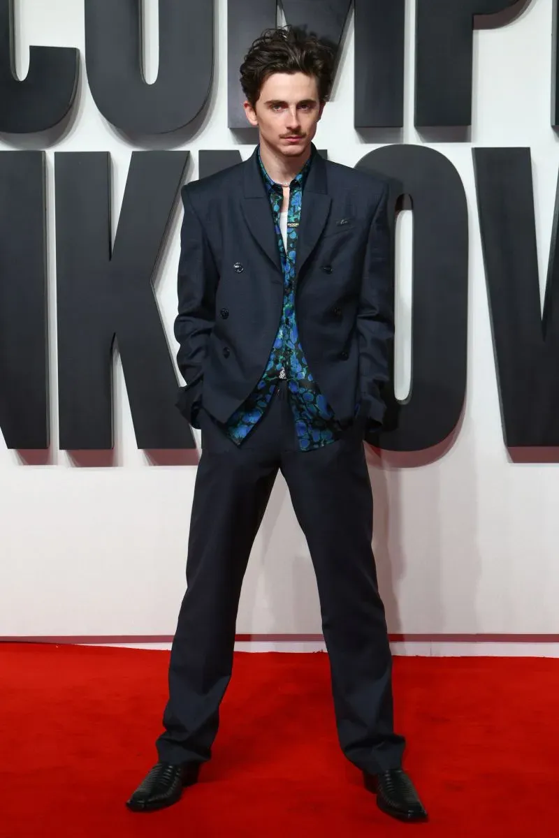 تيموثي شالاميت يقف على السجادة الحمراء بفيلم \"A Complete Unknown\"، مرتديًا بدلة مارتين روز مع قميص مزخرف، مع خلفية بارزة.