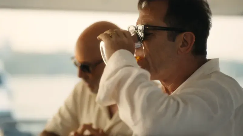 رجل يرتدي نظارات شمسية ويشرب مشروباً في مشهد من مسلسل "اللوتس الأبيض"، حيث تتناول القصة تأثيرات اللورازيبام على الشخصيات.