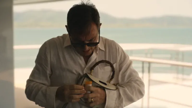 رجل يرتدي نظارات شمسية يفتح زجاجة دواء في مشهد من مسلسل "اللوتس الأبيض"، مما يشير إلى قلقه بشأن تأثيرات اللورازيبام.