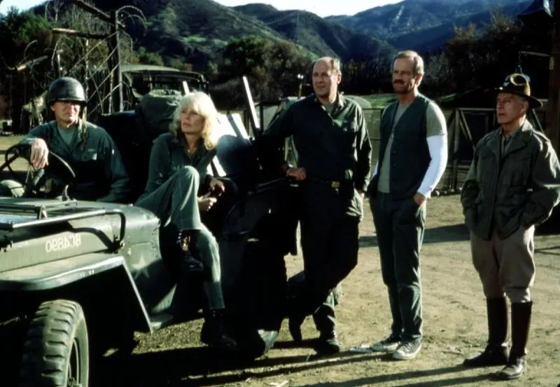 مجموعة من الشخصيات من مسلسل "M.A.S.H." يقفون بجانب سيارة جيب عسكرية في موقع تصوير، مع خلفية جبلية.