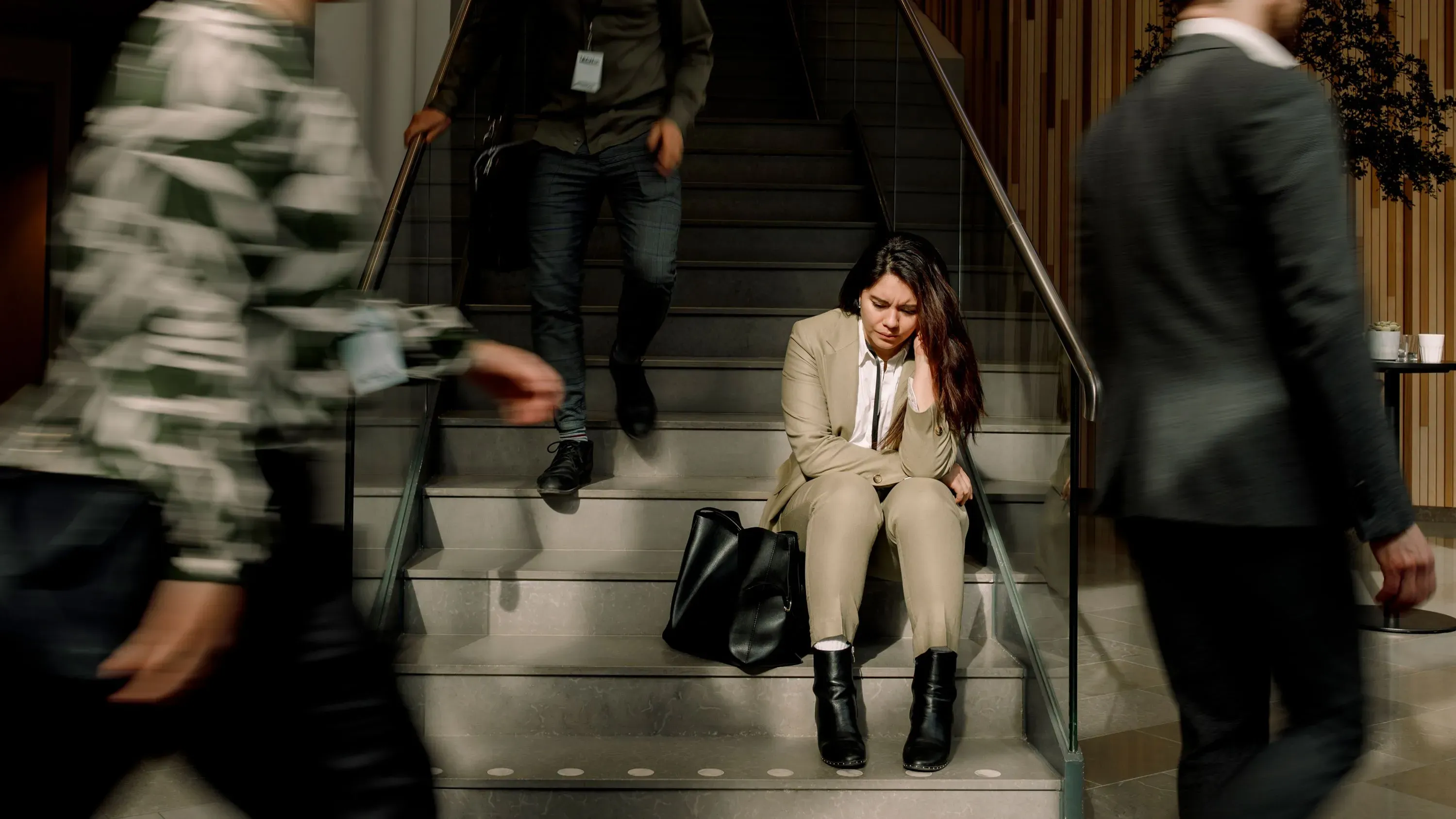 امرأة تجلس وحدها على الدرج، تبدو قلقة ومتوترة، بينما يمر من حولها أشخاص في بيئة مكتبية، مما يعكس تأثير فقدان الوظيفة على الصحة النفسية.