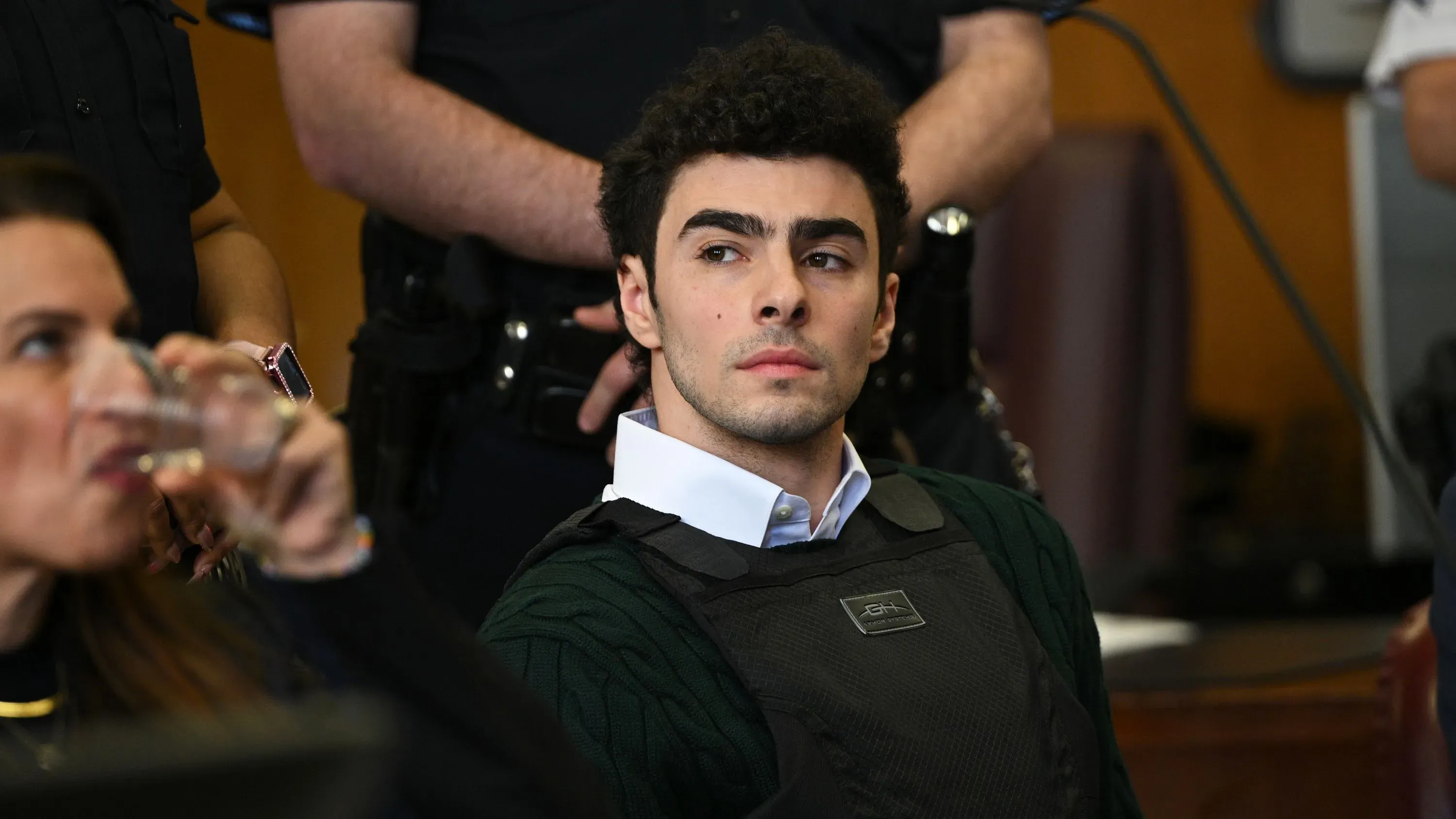 لويجي مانجيوني، المتهم بقتل الرئيس التنفيذي لشركة UnitedHealthcare، يجلس في قاعة المحكمة مرتديًا سترة واقية، مع محاميه في الخلفية.