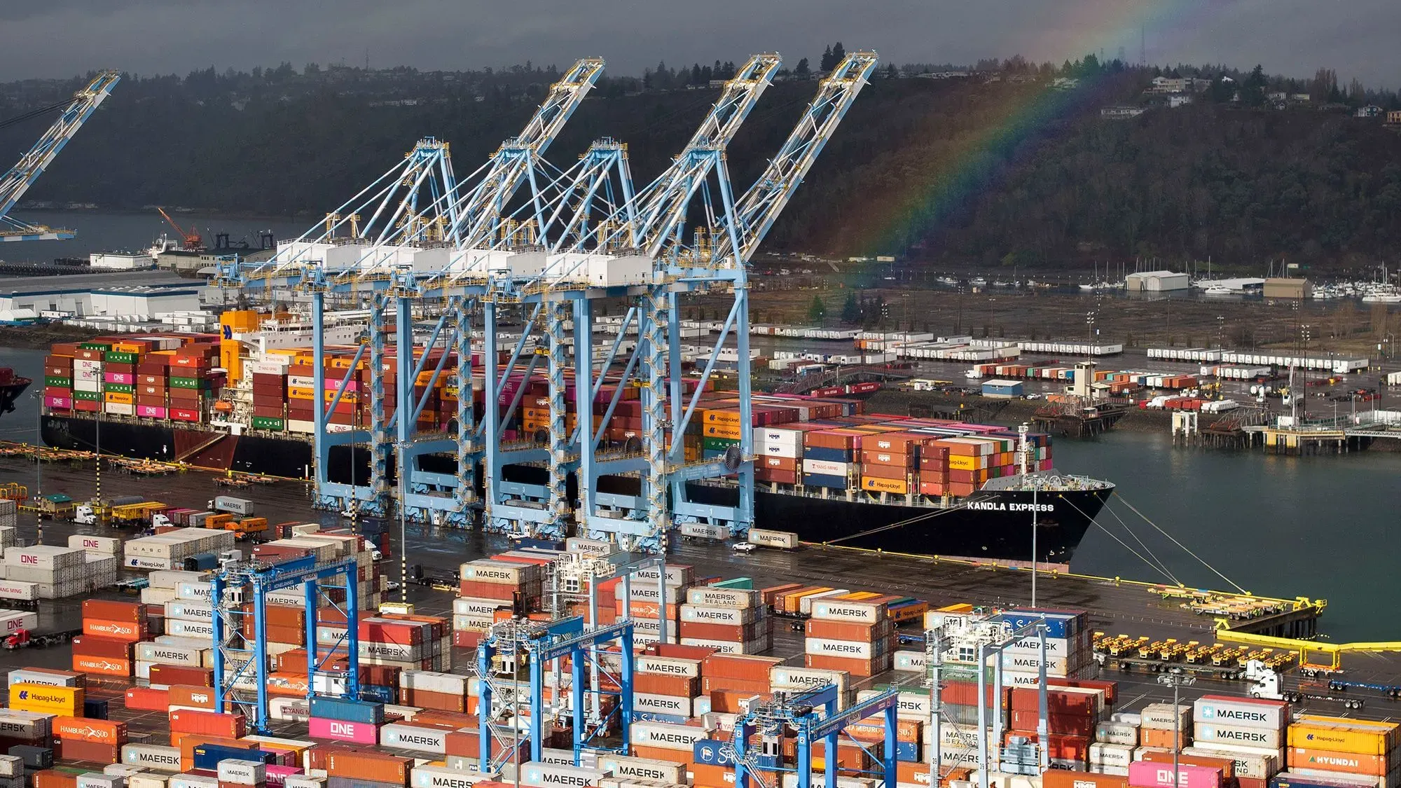 صورة لميناء يحتوي على حاويات شحن ملونة وسفينة "كانولا إكسبريس"، تعكس تأثيرات سياسة التعريفات الجمركية على التجارة.