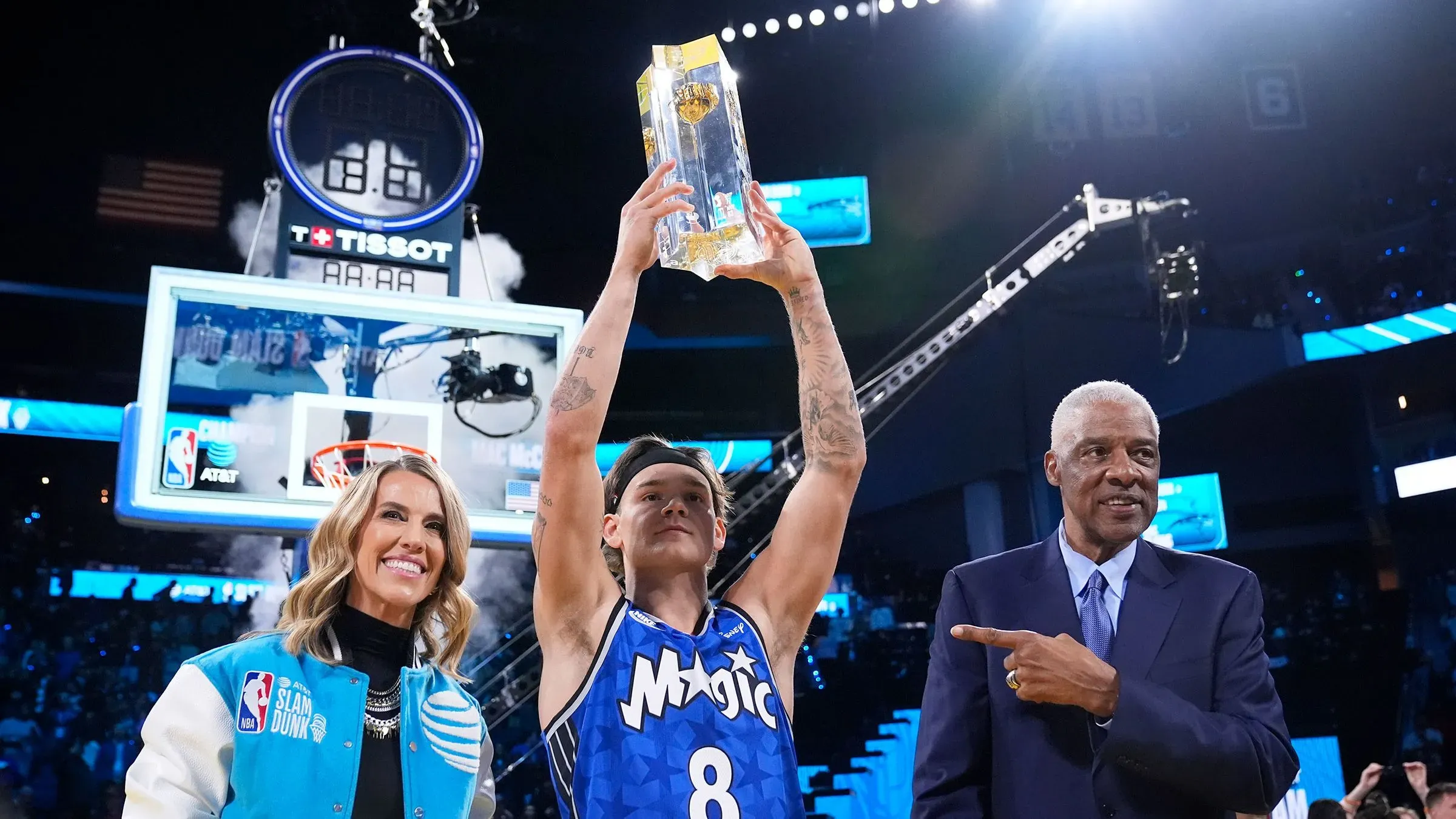 ماك ماكلونج يحمل الكأس بعد فوزه بمسابقة سلام دانك في الدوري الأمريكي للمحترفين، مع الابتسامة واحتفال الحضور في الخلفية.