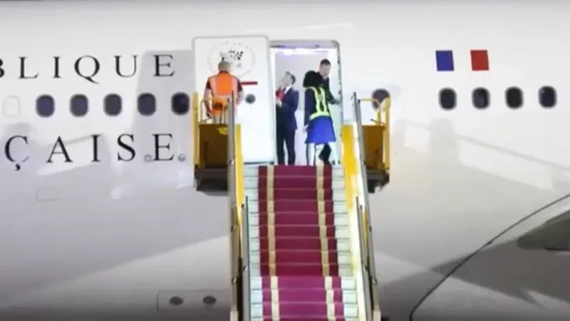 ماكرون وزوجته بريجيت ينزلان من الطائرة في فيتنام، مع مشهد للدرج الأحمر والموظفين في الخلفية، في لحظة غير رسمية.